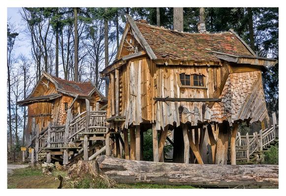 The picture shows two rustic tree houses built on wooden poles in a forest setting. The houses are made of weathered wood with scaly, roof tile-like cladding, which reinforces the impression of age and naturalness. The roofs are covered with moss-covered tiles and have antler-like decorations on the gables. Both houses have external staircases with wooden railings leading to the entrances and stand on high poles that raise them above the forest floor. Tall trees with thin, bare branches can be seen in the background. There are large tree trunks and some grass on the ground, emphasizing the natural surroundings. The light is soft, with a clear sky shimmering through the treetops.