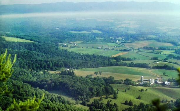 Looking out from a rock outcrop high atop a ridge crest down over the treetops to a farming valley hundreds of feet below. Beyond the wooded foothills, there are large green fields broken by strips of forest, rural lanes and farm buildings. There are also larger swaths of forest throughout the valley. In the misty distance another long, relatively flat-topped mountain range rises from the far side of the valley.