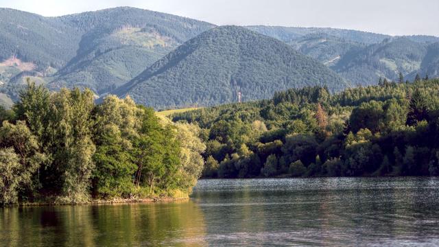 Evening photo looking across a lake to a forest on the hill on the opposite shore. There is a small tree-covered island in the middle, lit by the setting sun. Far in the background are higher mountains.
