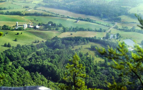 Looking out from a rock outcrop high atop a ridge crest down over the treetops to a farming valley hundreds of feet below. Beyond the wooded foothills, there are large green fields broken by strips of forest, rural lanes and farm buildings. Two small, roughly rectangular ponds that appear to be manmade reflect the hazy summer sky to our right.