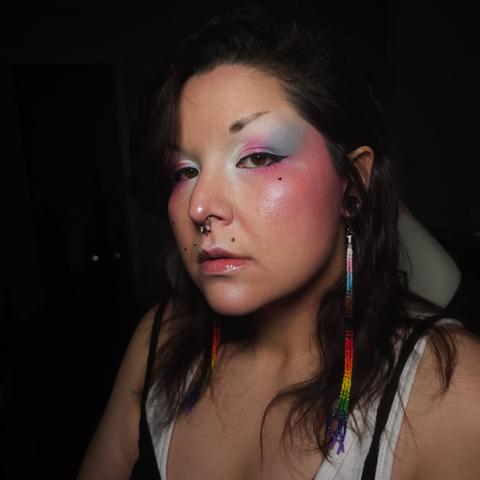 Ashley wearing a pair of long progress pride beaded dangle earrings and eyeshadow in the trans pride colors, from white at the inner corner of the eye, to pink in the middle of the lid, and blue at the outer corner.