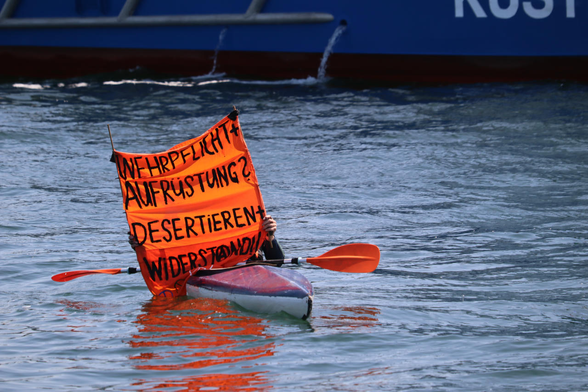 Zu sehen ist ein Kanu im Wasser. Darin sitzt ein Mensch welcher einen Orangenen Banner vor sich hält. Auf dem Banner steht "Wehrpflicht und Aufrüstung? Desertieren und Widerstand!"