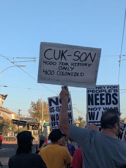 Black-on-white protest sign against a blue sky, held by a marcher. Text: cuk-son 4000 year history only 400 colonized.