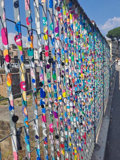 Photo of a long metal fence against the distant backdrop of Roman ruins. The fence is covered in circular stickers in all the colors of the rainbow, having a very cheerful appearance. The stickers are used by people who belong to walking tour groups.