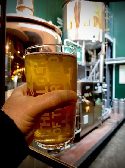 Hand holding a clear, golden colored beer in a glass with a copper-cladded brewhouse in the background.