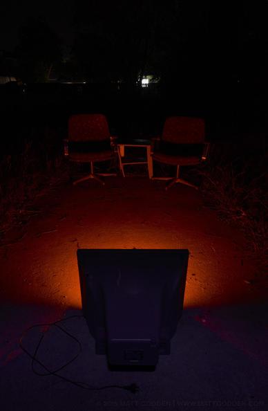 An image of an unplugged CRT television, facing away from the viewer. Orange-red light is flooding out of it, as if from an open fire. Illuminated in that light, are two matching discarded office chairs, with the broken remains of a small wooden coffee table between them. The whole tableaux appears as a post-apocalyptic scene. The background is all darkness, apart from the silhouettes of trees and fencing of the boundaries of this empty residential lot.