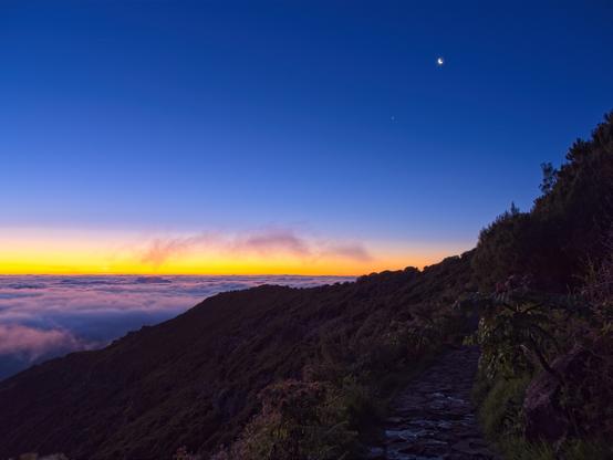 A crescent Moon and Venus hang in the upper right of a deep blue, pre-dawn sky above thick clouds. The horizon is yellow-orange as the sun approaches. The lower right quadrant of the image shows the dim terrain of the mountain path.
