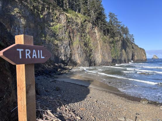 Wooden trail sign on a rocky beach with cliffs and waves in the background under a clear blue sky.