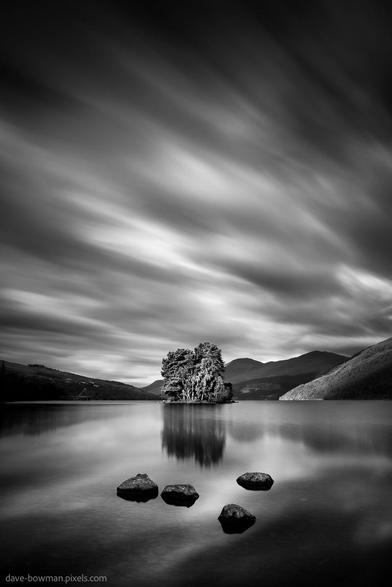 A photograph of four dark rocks emerging from the still surface of Loch Tay in the foreground, all rendered in monochrome. Behind them, the loch stretches out toward a small, tree-covered island near the centre of the frame. Above, streaks of cloud blur dramatically across the sky, their motion contrasting with the quiet water below. The black and white tones give the scene a timeless, otherworldly quality, emphasising both its calm and its subtle intensity.