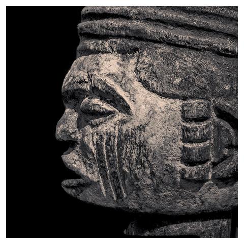 The picture shows a detailed, antique-looking sculpture of a human face in profile, made of wood. The sculpture has a rough, weathered surface with visible cracks and worn areas that indicate age. The head is decorated with an elaborate, tiered hair or headdress arranged in horizontal lines. The facial features are striking: closed eyes with deep wrinkles, a prominent nose and a slightly open mouth. Vertical grooves or scars are engraved on the cheek, looking like ritual markings. The background is completely black, which highlights the sculpture and gives it a dramatic contrast. The color scheme is in monochromatic tones, mainly shades of grey and brown.