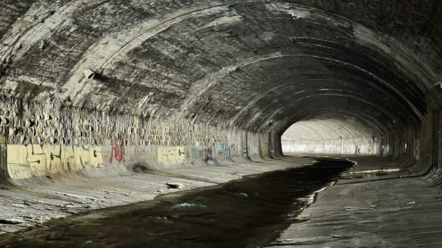 Ein dunkles, altes Gewölbe, etwa 10 Meter breit und 6 Meter hoch. In der Mitte fließt ein Fluss, aktuell mit niedrigem Wasserstand. Links und rechts ein leicht abschüssiges Ufer. Im Hintergrund eine Kurve, hinter der es heller zu werden scheint. An den Wänden etwas Graffiti.