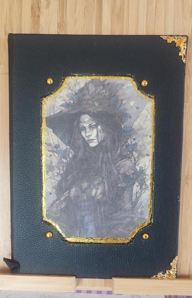 Blakc cover, with a priunt of a withc with blakc robe in the centre, the frame is embossed, and on the four corner of the pictures there  are decorative rivets. There is gold leaf decoratio around the print. On the two book corners there are some gold decoration