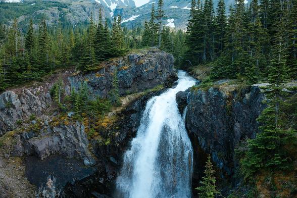 A waterfall, front and centre. It falls into a rocky faced valley. The top of the falls are lined with evergreen trees and there are mountains in the distance.