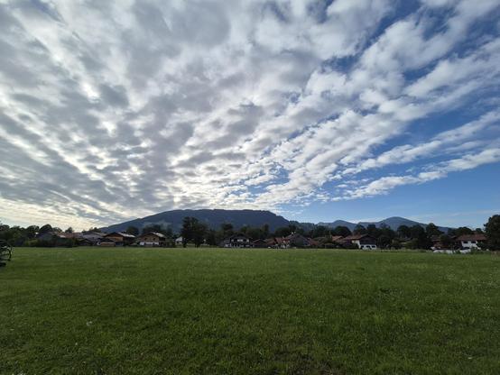 A green field with houses and mountains in the back. A blue sky with white clouds on top