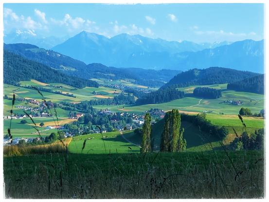 A village is in a valley, surrounded by hills with forest. In the background a range of mountains in the haze.