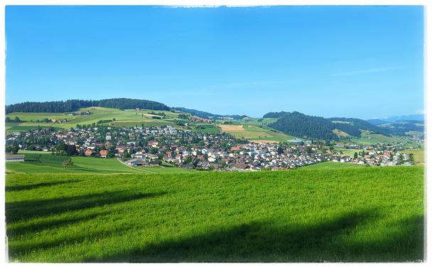 Behind a green field is a village at the slope of a hill. Blue sky.