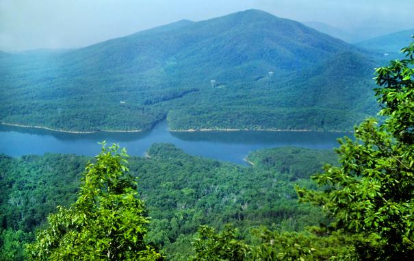We are high atop a ridge crest, looking down over the treetops down into a valley hundreds of feet below with a long narrow lake wrapping around a small wooded mountain on its far shore. The entire view is densely forested except for a single power transmission line. Barely visible through the haze beyond is another wooded mountain. The hazy sky is a very soft, washed-out light blue.