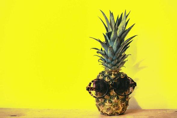 A pineapple sitting in front of a bright yellow wall. It has a pair of sunglasses hanging on the fruit as if the pineapple has eyes. The green leaves of the pineapple are still attached to it and look like a spiky hairdo.