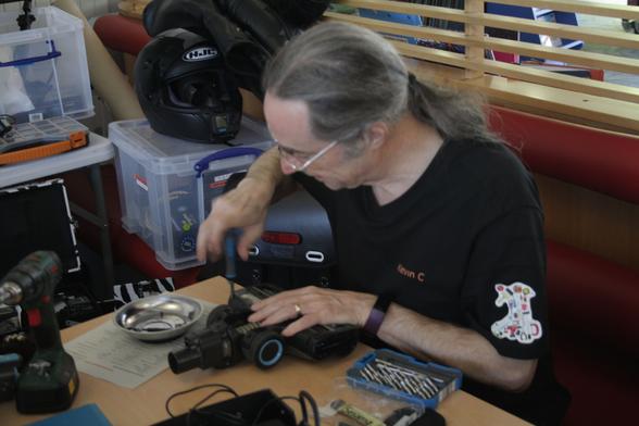 Kevin, a man with light skin tone and long grey hair tied back, sat at a table working on the head of a vacuum cleaner. On the table are various screwdriver heads, a silver bowl with screws in it, an electric screwdriver. He is holding a manual screwdriver and using it on the head.