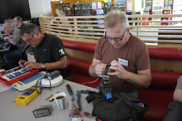Kevin, a man with tanned light skin tone and short black hair, and Gary, a man with light skin tone and short blonde hair, sitting a table. Kevin is working on a red toy musical keyboard while Gary is working on what appears to be part of a vacuum cleaner. On the table is a mug of coffee, various electrical sockets, and a screwdriver.