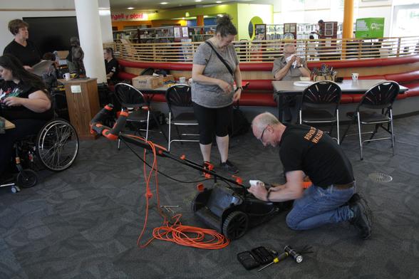 Richard, a man with light skin tone and short grey hair, kneeling on the floor working on a lawn mower while a woman with light skin tone and long brown hair tied up in a bun stands watching. Around them are tables at which different people are sitting working on items.