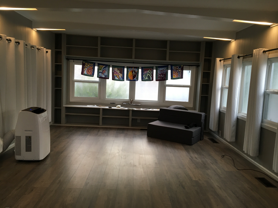 A nearly empty living room , apart from an portable air conditioner, some prayer flags, and a foam chair thing.