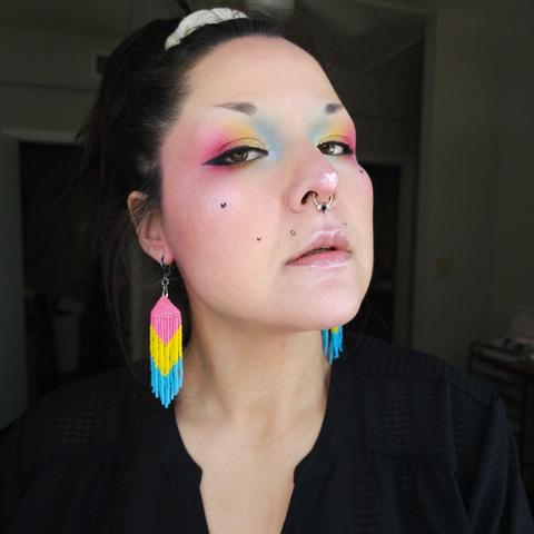 Ashley wearing a pair of pansexual pride beaded fringe earrings and eyeshadow in the pansexual pride colors, from blue at the inner corner of the eye, to yellow in the middle of the lid, and pink at the outer corner.