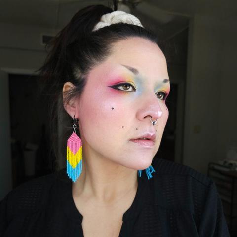 Ashley wearing a pair of pansexual pride beaded fringe earrings and eyeshadow in the pansexual pride colors, from blue at the inner corner of the eye, to yellow in the middle of the lid, and pink at the outer corner.