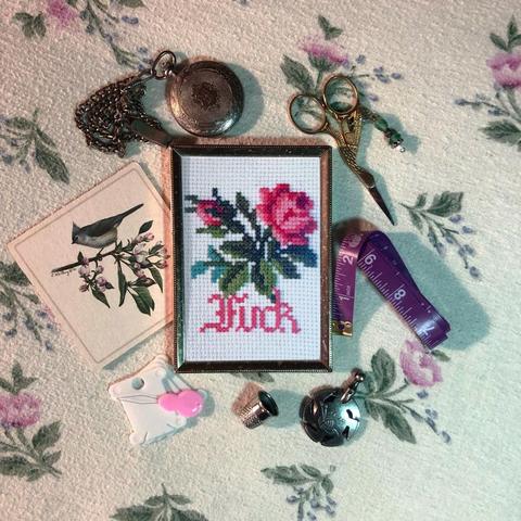a small piece of cross stitch inside a thin gold frame surrounded by various bits of vintage looking sewing accouterments. the cross stitch is of a small pink rose and bud with bright green leaves. under the rose is the word "Fuck" in matching pink calligraphy.