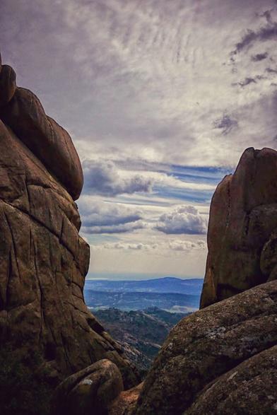 A dramatic view between two large, weathered rock formations that frame a distant mountainous landscape under a cloudy sky. The lower part of the scene shows a hazy, bluish valley with rolling hills, suggesting a vast distance. The sky above is filled with a mix of fluffy white and darker grey clouds, creating a sense of depth and atmosphere.