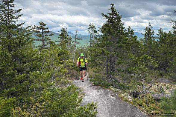 A hiker wearing a bright green shirt, black shorts, and a green cap treks along a narrow, rocky trail in a dense evergreen forest. They carry a backpack and use trekking poles for support. Towering trees frame the path, and distant mountains rise under a cloudy sky, adding depth to the landscape.