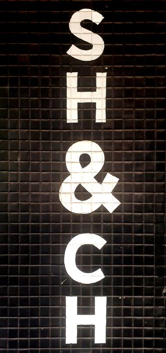 A section of a shop sign painted onto a black ceramic tiled wall. The letters vertically read SH & CH