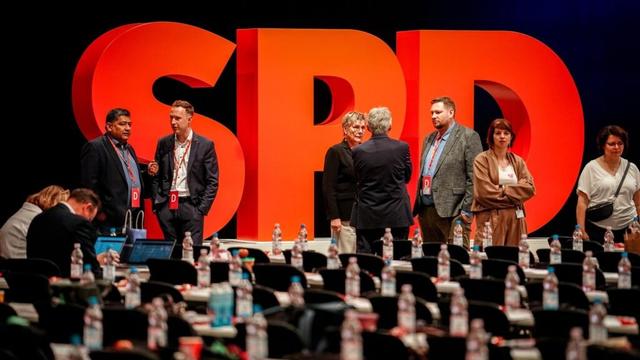 Auf der Bühne vor einem SPD-Schriftzug aus stehenden großen roten Buchstaben stehen mehrere Teilnehmer und reden. Im Vordergrund unscharf die Delegiertenbänke.