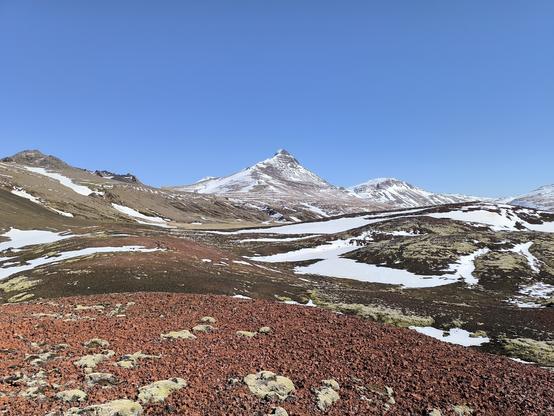 Une zone de roches volcaniques rouges au premier plan. Plus moins, d'autres zones volcaniques recouvertes de mousse vert pâle et partiellement enneigées. Au fond, une montagne au sommet pointu se détache du paysage. Le ciel est totalement dégagé.