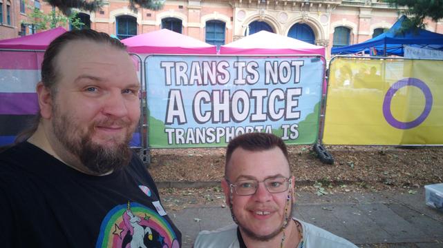 Two white guys are standing in front of a banner that says "trans is not a choice, transphobia is". I'm the tall fat blond guy off to one side, E is the short guy in front.