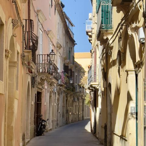 Narrow sunlit alleyway in a historic European town, lined with tall, closely packed buildings featuring ornate balconies, shuttered windows, and pastel-colored facades. A motorbike is parked along one wall, and the street curves gently in the distance under a clear blue sky.