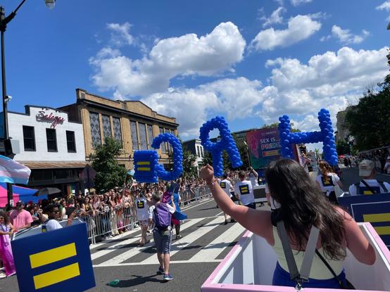 The view of the World Pride parade in Washington DC, from the point of view of a person in the parade, walking with HRC.  Large letters formed from balloons (spelling PRIDE), as well as people in boxes with the HRC equal-sign logo, are seen in the street; large numbers of people line the sidewalks.