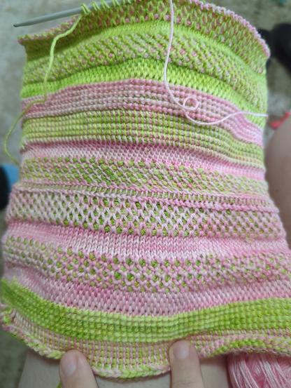 A Tunisian crocheted fabric draped over (presumably) a knee. It's still in progress, the double ended hook can be seen at the top holding some live stitches. it's a cylinder laid flat. It's made in 2 (slightly variable) colors, the working ends of each are visibly trailing off from either end of the hook. One color is a nice spring green and the other is a lovely spring pink. The entire thing is worked in bands of wildly different stitches, each roughly an inch thick. Each band of stitches looks totally different, and uses the 2 colors in different ways. Some bands only have one or the other color visible, sone have both but all in very different ways. Tiny bits of a person's leg and hand can be seen at the very periphery of the photo.