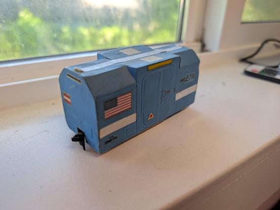 Old concept model I built in 1979 for a boxcar to be used on a lunar railroad. It is cardstock and 1/48th scale. It is light blue, with various stripes and small decals, boxy, short, and low to the ground. There are two sets of wheels, but they aren't visible in the photo. The sides of the roof are beveled, except for a plain-box center section with double pressure loading doors on each side. 
This high side-quarter view has a large American flag to the left of the door.