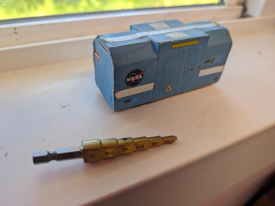 Old concept model I built in 1979 for a boxcar to be used on a lunar railroad. It is cardstock and 1/48th scale. It is light blue, with various stripes and small decals, boxy, short, and low to the ground. There are two sets of wheels, but they aren't visible in the photo. The sides of the roof are beveled, except for a plain-box center section with double pressure loading doors on each side. 
  This is the reverse side from the previous photo, and while mostly identical, has a classic blue-circle NASA "meatball" logo to the left of the loading doors. A step drill bit sits in the foreground for scale. The model is about 4" long.