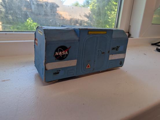 Old concept model I built in 1979 for a boxcar to be used on a lunar railroad. It is cardstock and 1/48th scale. It is light blue, with various stripes and small decals, boxy, short, and low to the ground. There are two sets of wheels, but they aren't visible in the photo. The sides of the roof are beveled, except for a plain-box center section with double pressure loading doors on each side.
