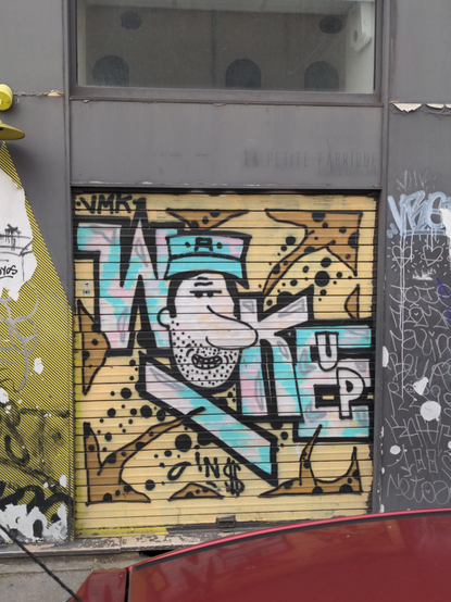 Sur un rideau métallique carré, sur fond jaune et entouré de flèches et de triangles motif guépard, le blaze "Wake Up" en blanc et en bleu en diagonale, le "A" étant remplacé par le visage allongé d'un type mal rasé avec un chapeau portant la lettre "A".
