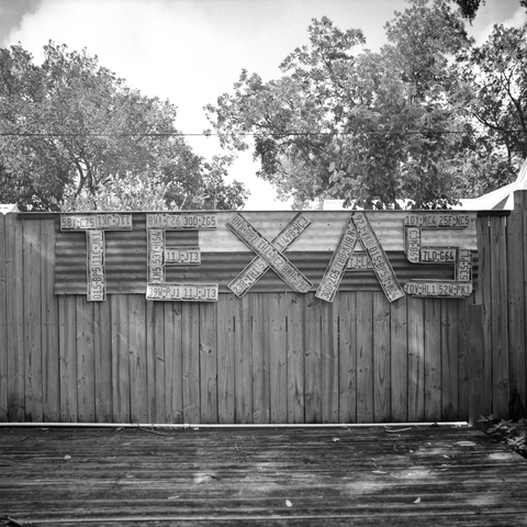 A black and white film photo. At Huebel’s Bier Garden, “TEXAS” is spelt on a wood fence using old license plates in Smithville, Texas. Sunday, June 29, 2025.