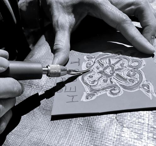 A close-up, black and white image shows a pair of hands using a stylus to carve a stamp block. The block features an intricate floral design with a central heart shape emerging. The stylus, held in a grip, is pointed toward the block where carvings are being made. The background consists of a textured surface that appears to be a workbench or crafting area on top of a tarp. The letters "HE..." and "T..." have already been cut onto the top left corner of the carve.