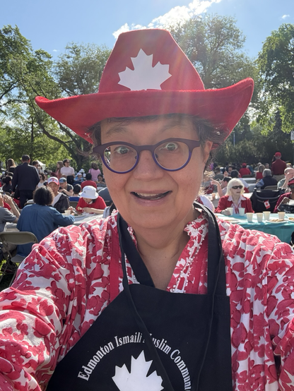 Senator Simons, in a red Canada Day cowboy hat, a red and white floral blouse, and a black and white apron, stands in front of rows and rows of tables, where a happy multicultural crowd, many in Canada Day attire, eat plates of pancakes on the sunny, leafy Alberta Legislature lower grounds.