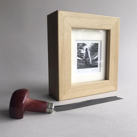 Photo of a small wood-effect frame on a plain background, with a wood engraving tool in the foreground. In the frame is a black and white print showing a standing stone in the centre, with circular rock art features. Grass is at the foot of the stone. The sky whirls around in the background.