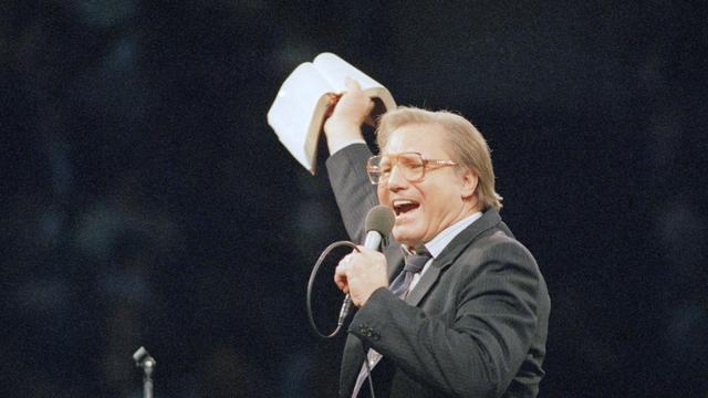 Jimmy Swaggart, before an audience, speaks into a microphone while holding a Bible overhead