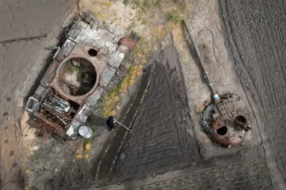 An Ukrainian man is raking a garden bed for sunflowers between two rusted tanks.
