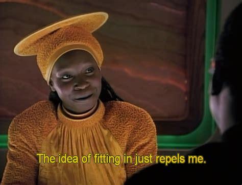Guinan, a character from Star Trek TNG, played by Whoopi Goldberg. She is wearing a yellow tunic and a headpiece that is tight to the head with a large disc on top. It is also yellow. There is a green frame behind her with what looks like part of an alien planet. She is talking to someone barely visible on the right, saying "The idea of fitting in just repels me." This is captioned under her in yellow writing.