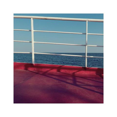 looking straight at the crooked angle of a ship's railing. the sky is blue, the ocean is bluer, the deck is red. the railing itself is casting some light morning shadows